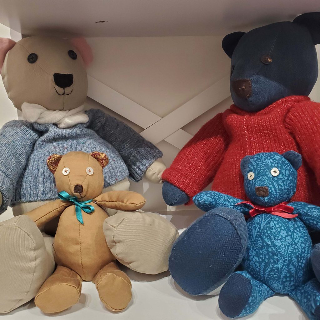 Photograph of handmade bears together on a shelf. The larger two bears at the back are wearing jumpers and the smaller two at the front have ribbons around their necks.